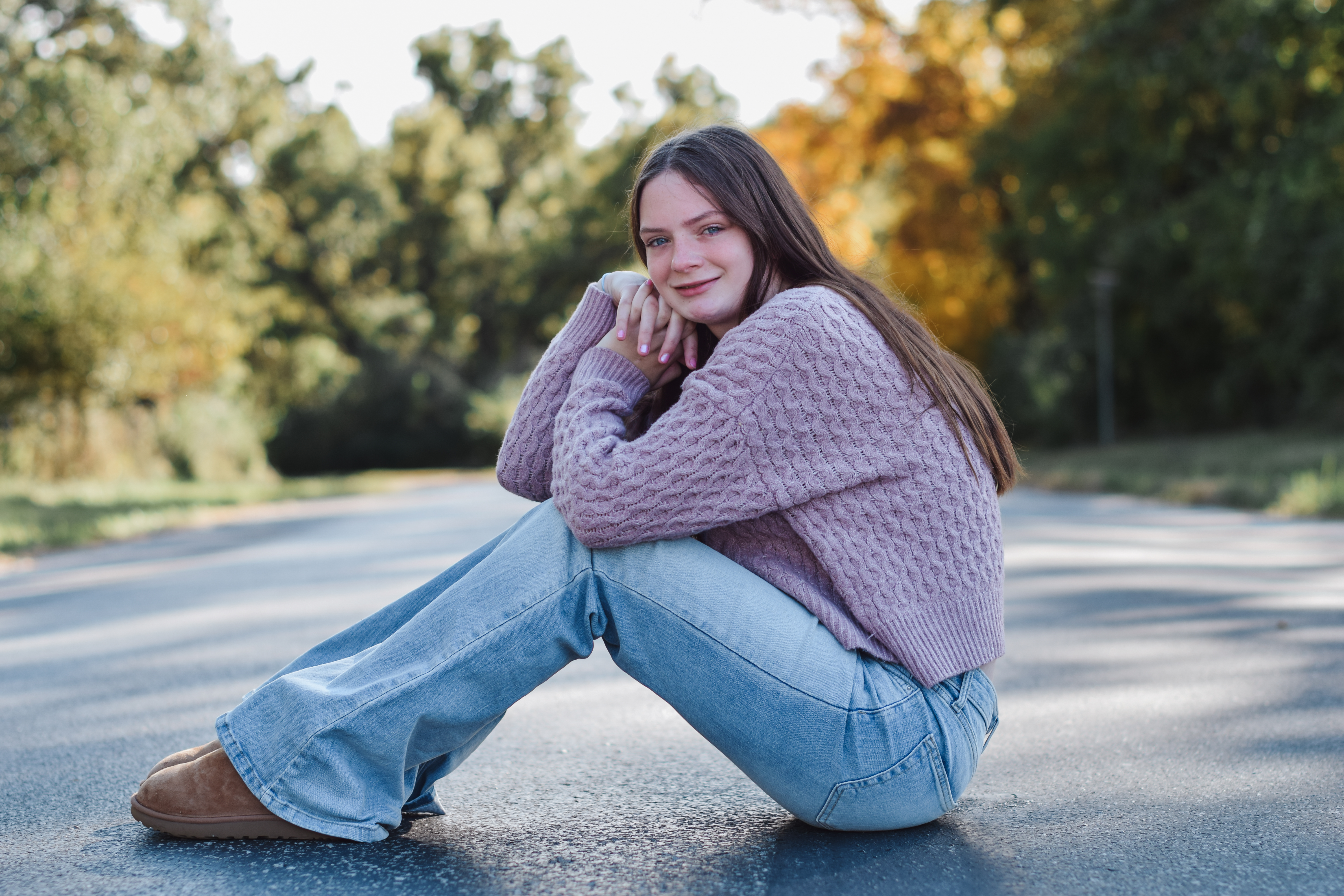 A young woman with long brown hair sits on the ground of a paved road, surrounded by trees in the background.