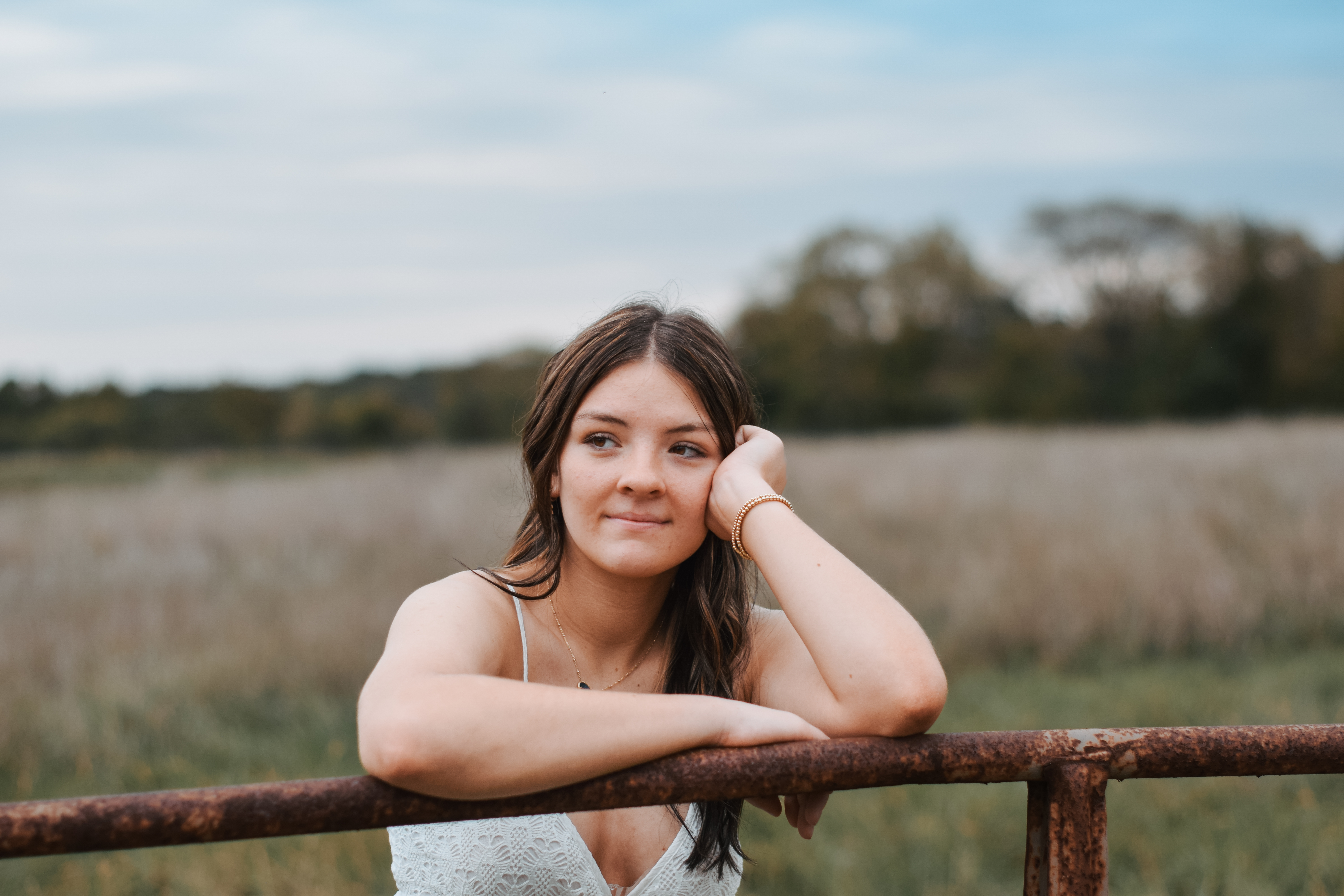 A young woman with long brown hair, wearing a white lace top, leaning on a rusty metal fence in a field.