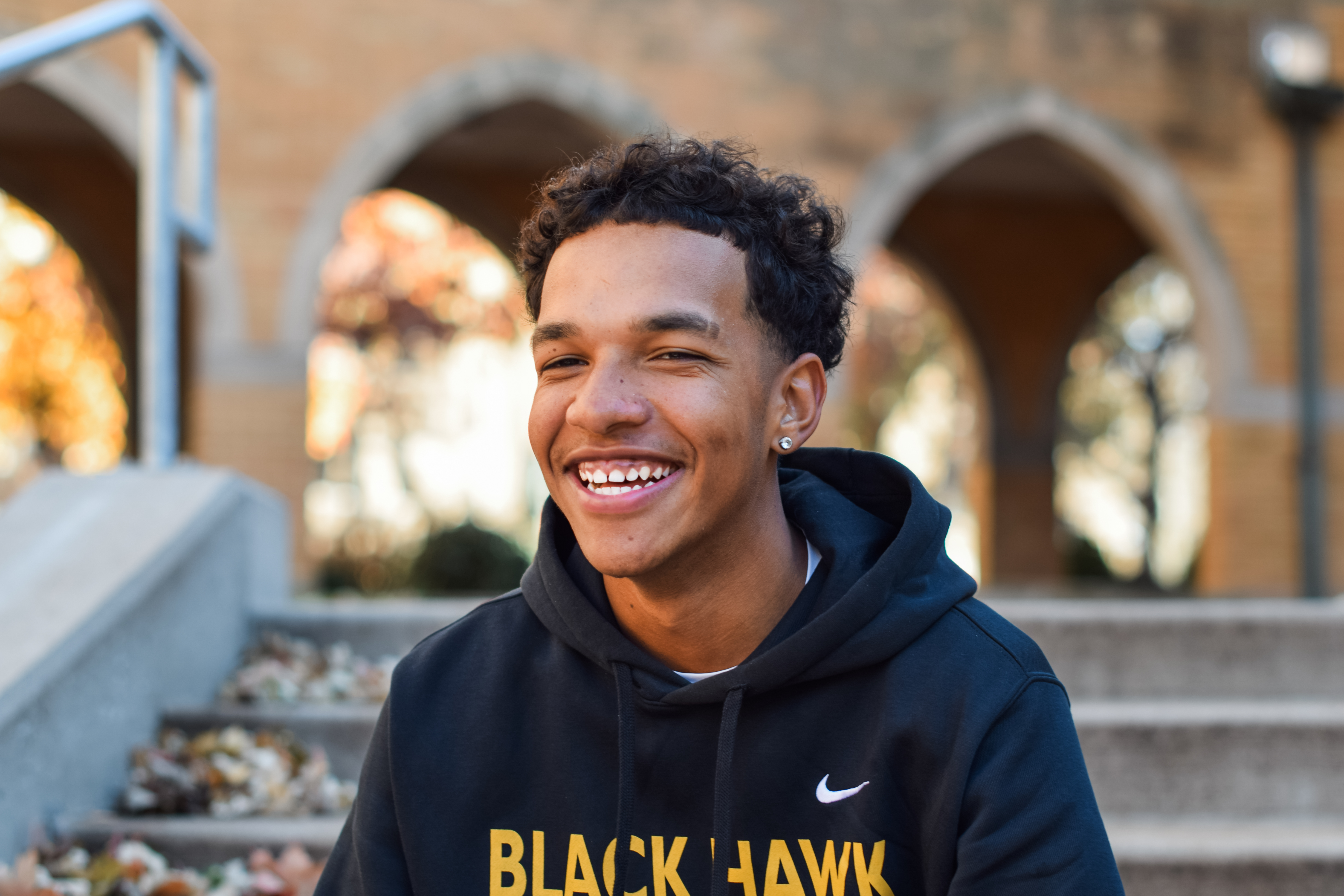 A young man with curly dark hair, wearing a black hoodie with yellow lettering and a white Nike swoosh, stands in front of...