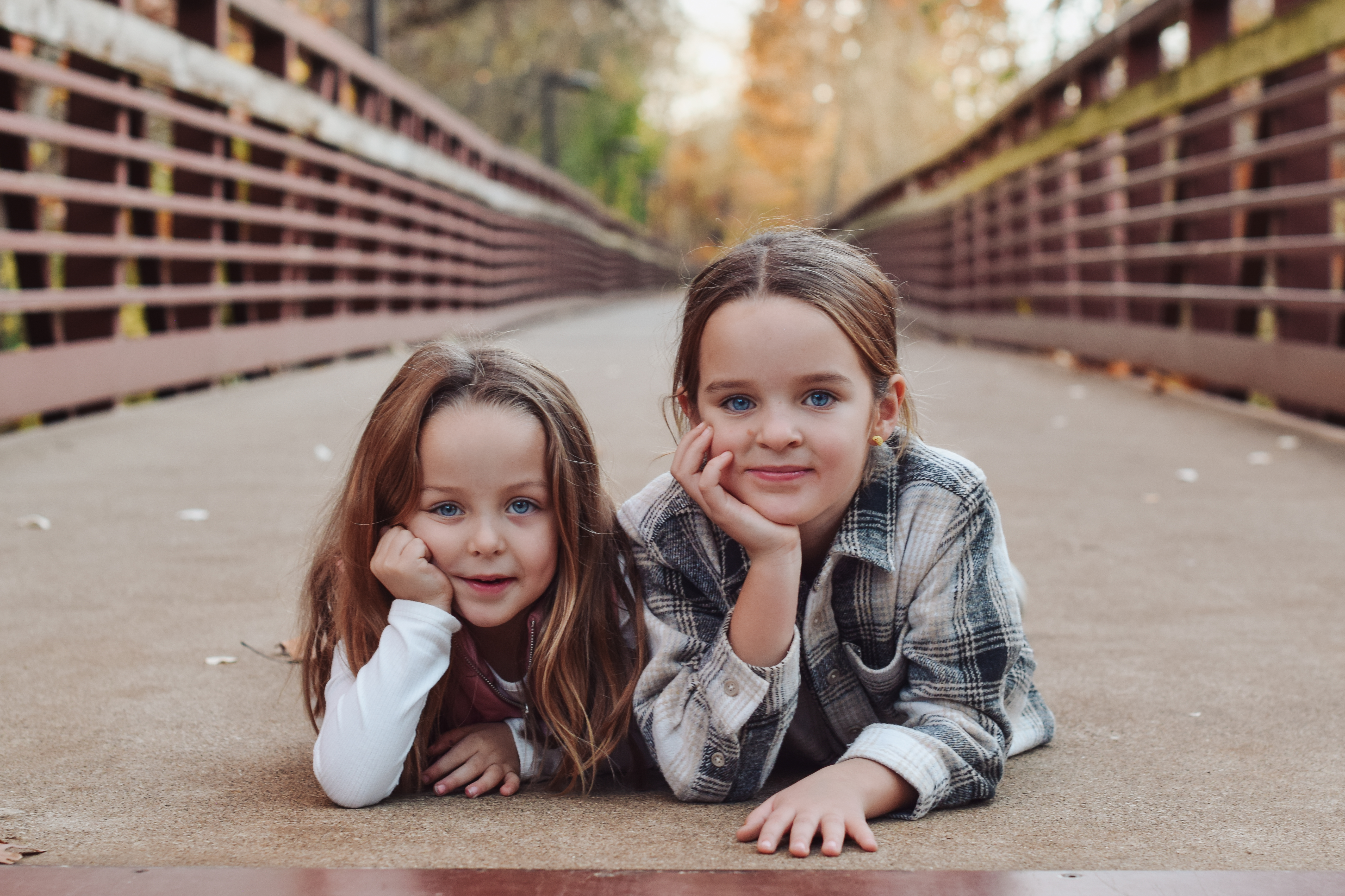 Two young girls laying on a bridge with their hands to their faces, smiling at the camera.